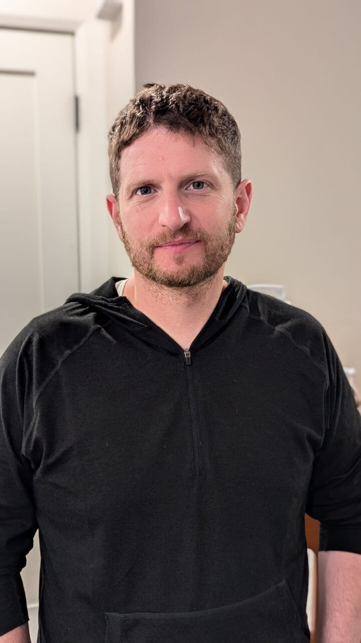 Photo of Merrick Levene, Merrick stands in front of a light background with a black V-necked shirt, with pale skin, short brown hair, dark eyes and a closely trimmed beard 